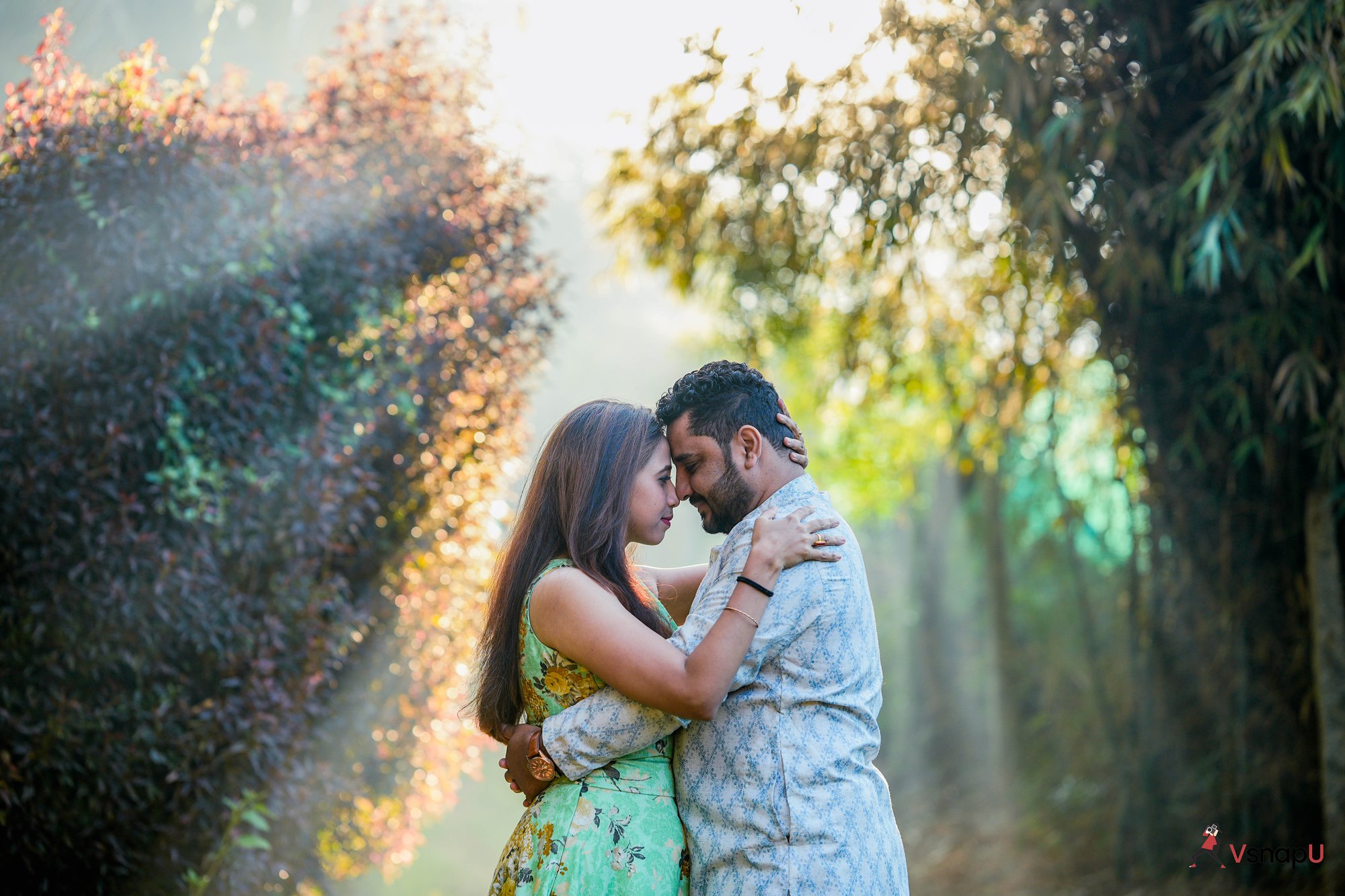 Pre-wedding photo of an ethnic couple in the forest, hugging with their heads touching as sunlight creates a serene atmosphere.